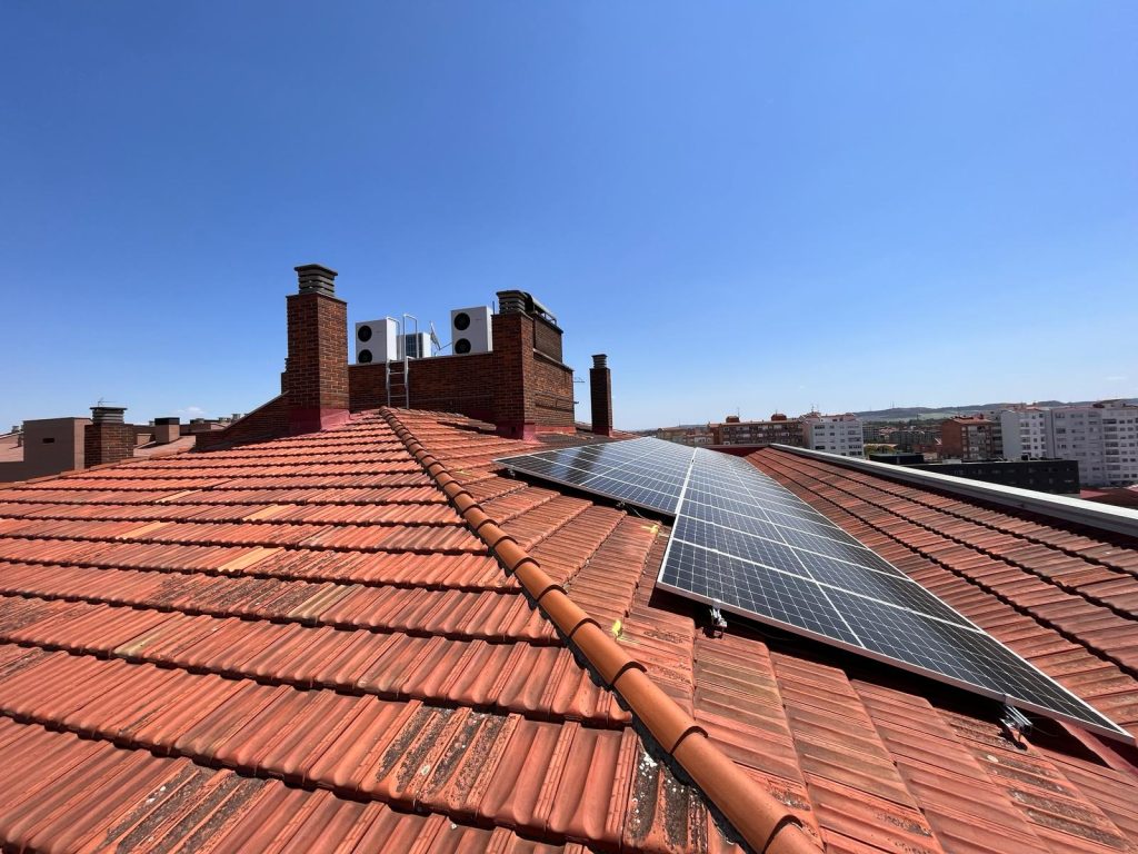 Paneles solares instalados sobre tejado de tejas rojas junto a unidades exteriores de bomba de calor aerotérmica en un edificio residencial, con cielo despejado y vista urbana al fondo.
