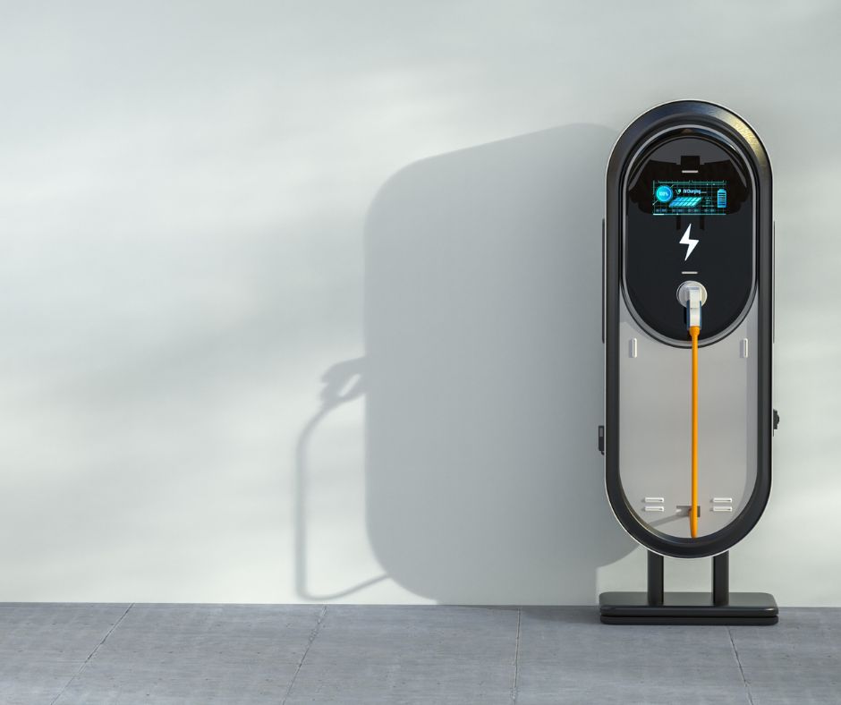 Cargador eléctrico vertical de diseño moderno con pantalla digital y cable naranja conectado, instalado junto a una pared gris clara sobre pavimento urbano.